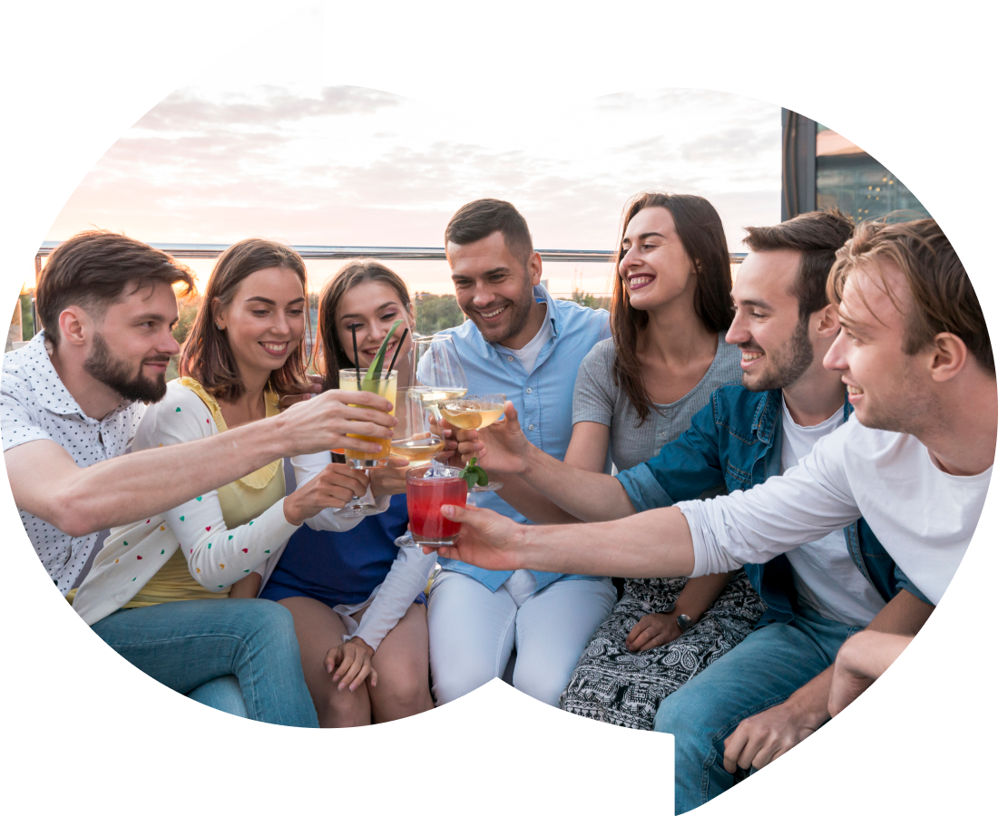 A group of seven friends sitting closely together outdoors, raising their glasses in a toast and smiling. They appear to be enjoying a casual gathering at sunset, with a view of the sky and surrounding scenery in the background.