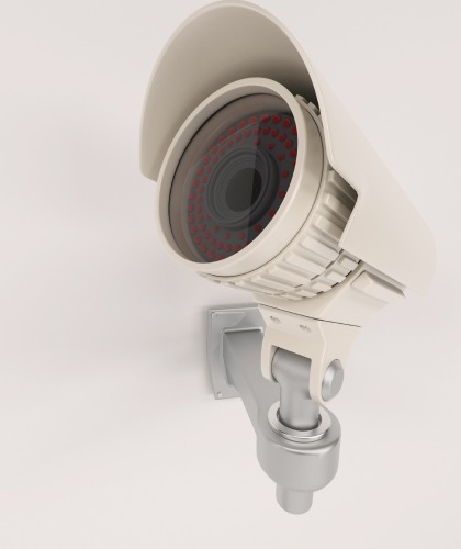 Стационарная охранная видеокамера в защитном кожухе, закреплённая на настенном кронштейне, направлена вперёд для видеонаблюдения.