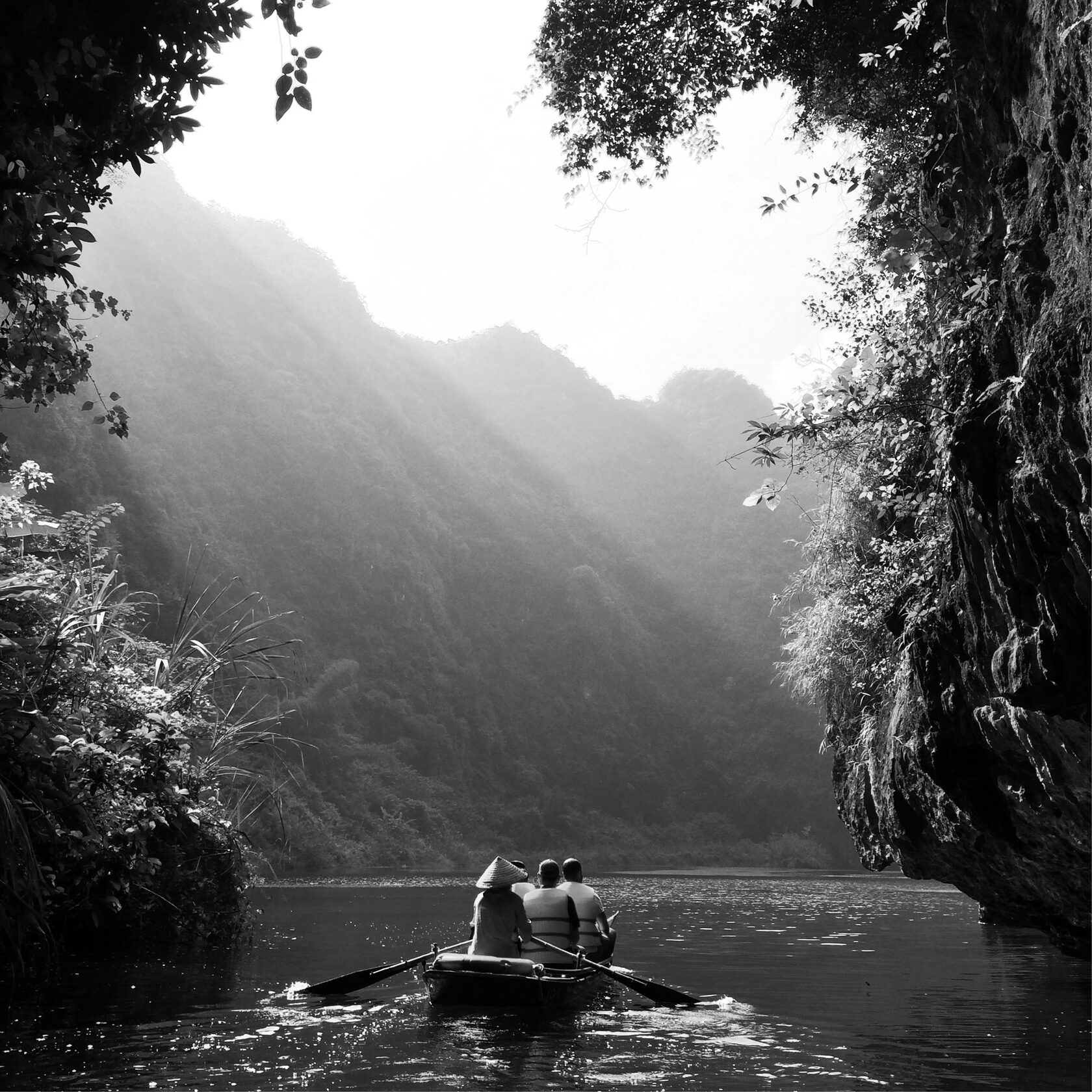 Чёрно-белая фотография гигантской статуи бога Муругана у входа в пещеры Батур (Batu Caves) в Малайзии. На заднем плане видна лестница, ведущая к пещерам, окружённая густой растительностью и скальным массивом. Монументальная фигура статуи доминирует в композиции, подчёркивая религиозное и культурное значение этого места. 