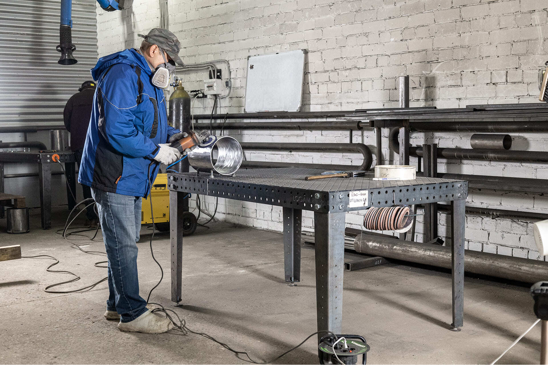 Сотрудник в защитной маске и спецодежде работает с металлической деталью вакуумной камеры на сварочном столе в цехе.