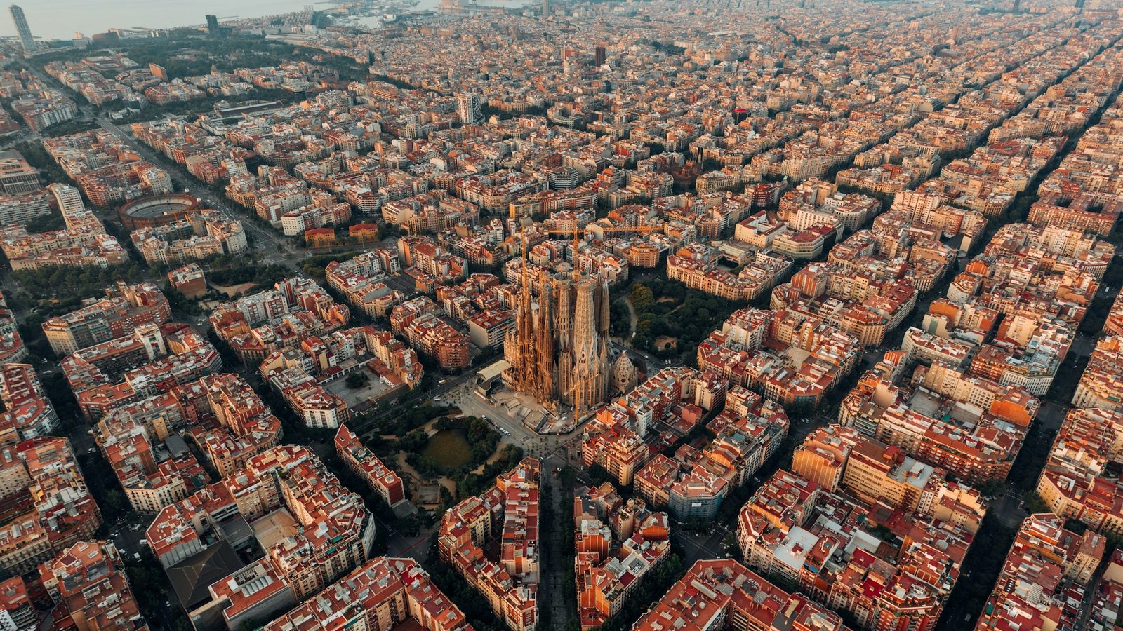 Aerial view of Barcelona's Eixample district at sunrise, with the Sagrada Família rising among the square city blocks and the sea in the distance