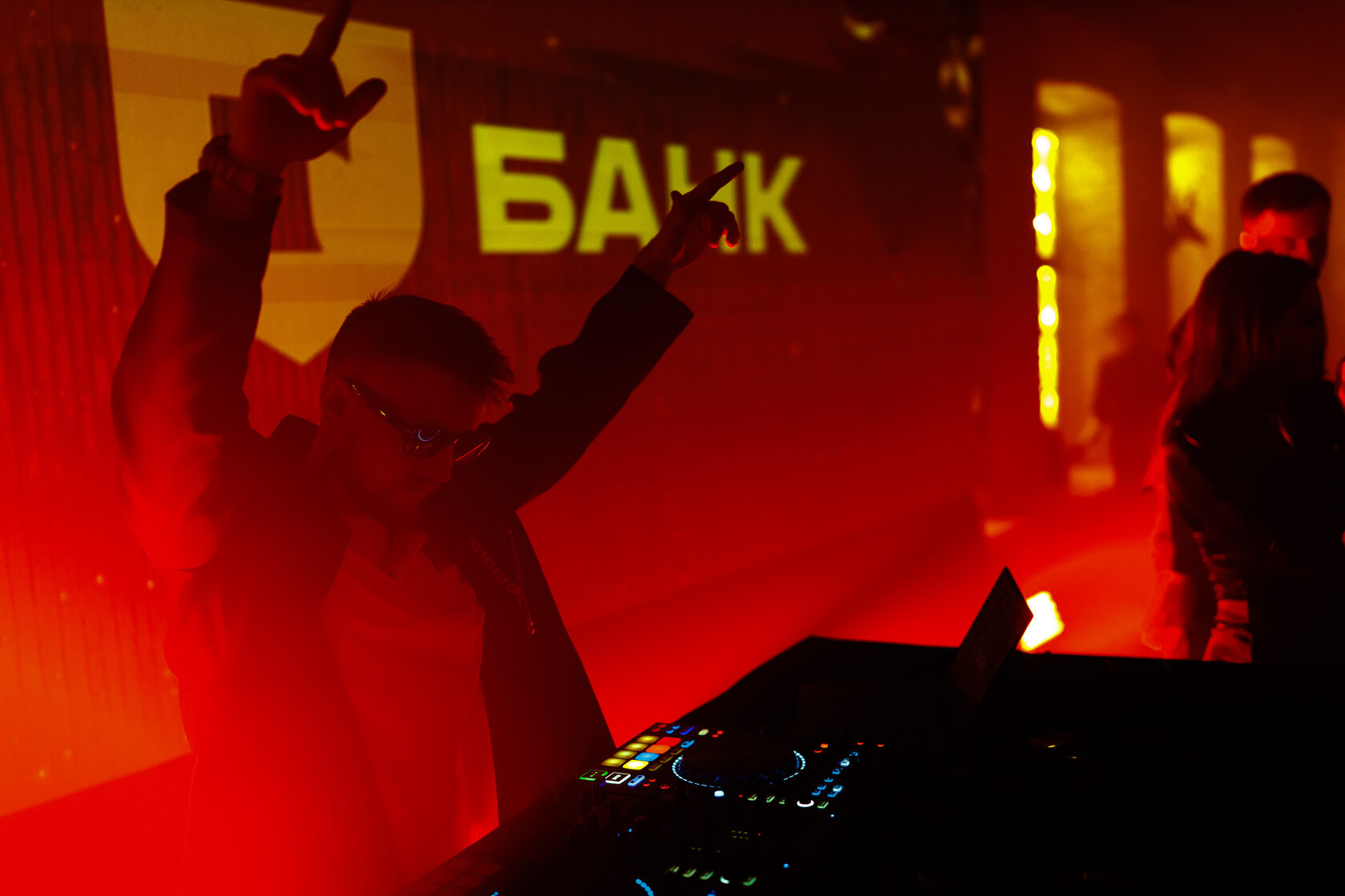 Силуэт DJ ROZHKOV с поднятыми руками за пультом в красном свете перед экраном с надписью банка.