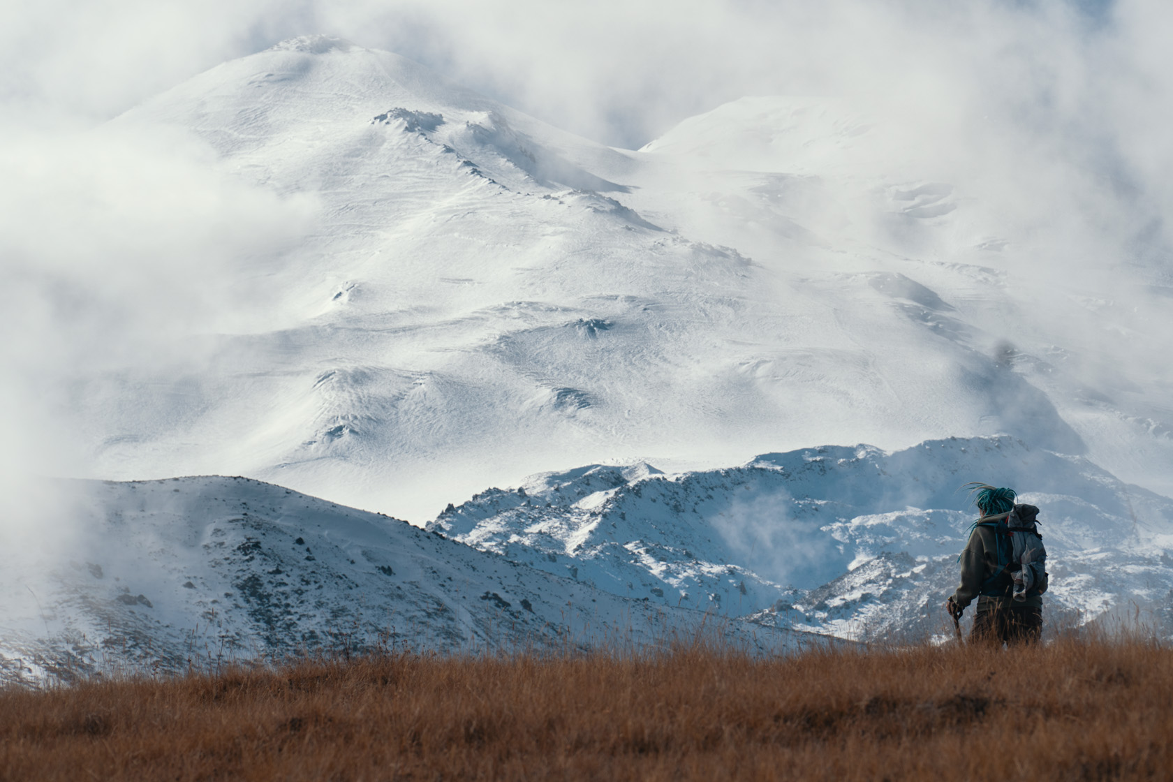 Северный склон горы Эльбрус. Гору Эльбрус затягивает облаками. На фоне горы Эльбрус идет девушка с рюкзаком. Фотограф Севальнев Ян.