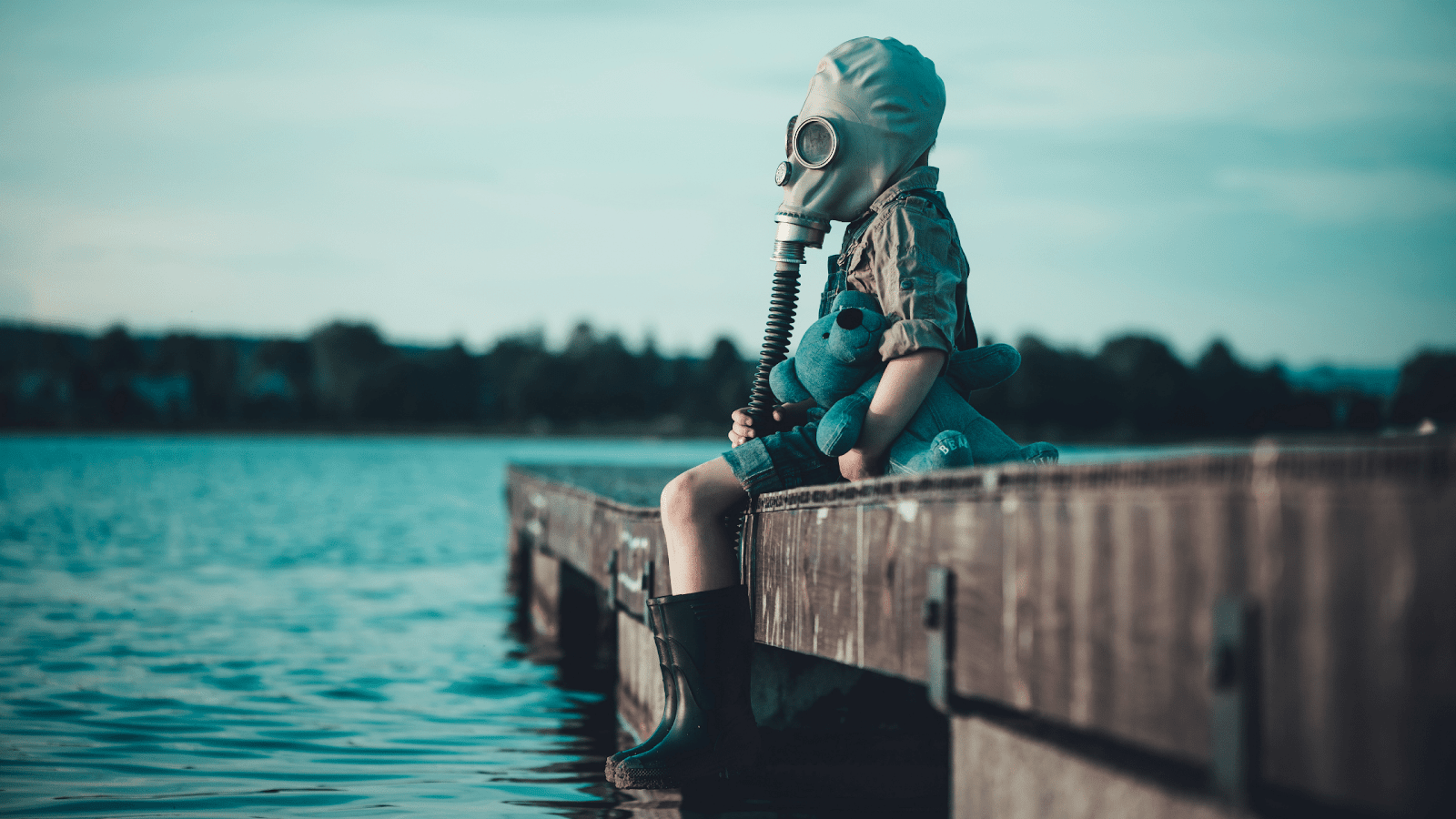 The child in the gas mask is sitting on the platform at the lake. Environment pollution.