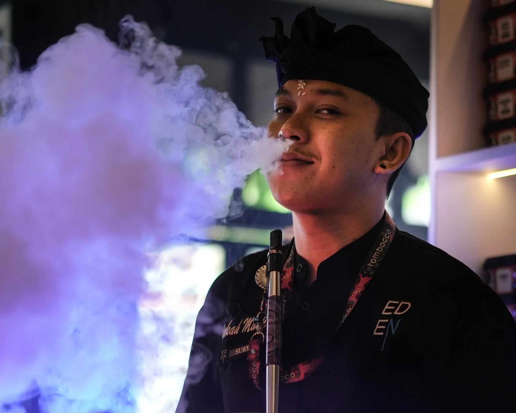 An expert shisha master in traditional Balinese attire exhaling a thick cloud of smoke at Eden Hookah Club.