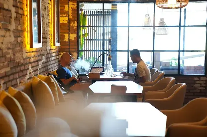 The warm and inviting interior of Eden Hookah Club, featuring exposed brick walls, soft yellow lighting, and large industrial-style windows. A couple of customers are seated at tables relaxing with hookahs, highlighting the venue's cozy atmosphere for evening networking.