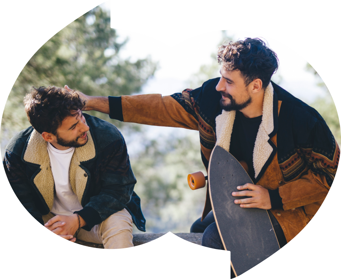 Two young men sitting outdoors, with one holding a skateboard. The man on the right is gently placing his hand on the shoulder of the other, who is looking at him and smiling. They are wearing casual jackets with sherpa linings, and the background shows a