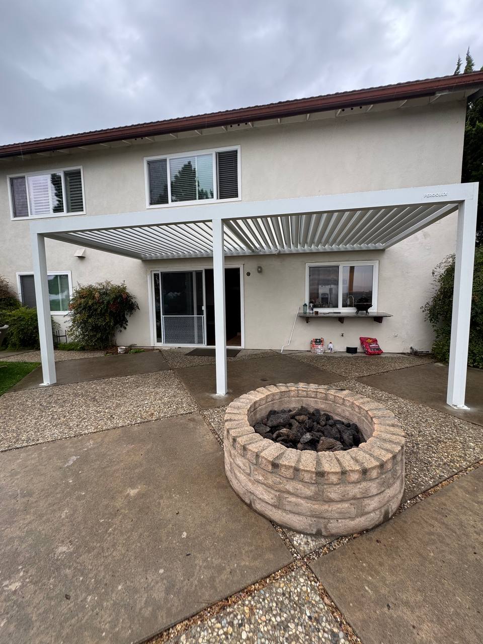 Completed white 10 by 19 foot wall-mounted motorized pergola in a Fountain Valley backyard beside a round fire pit