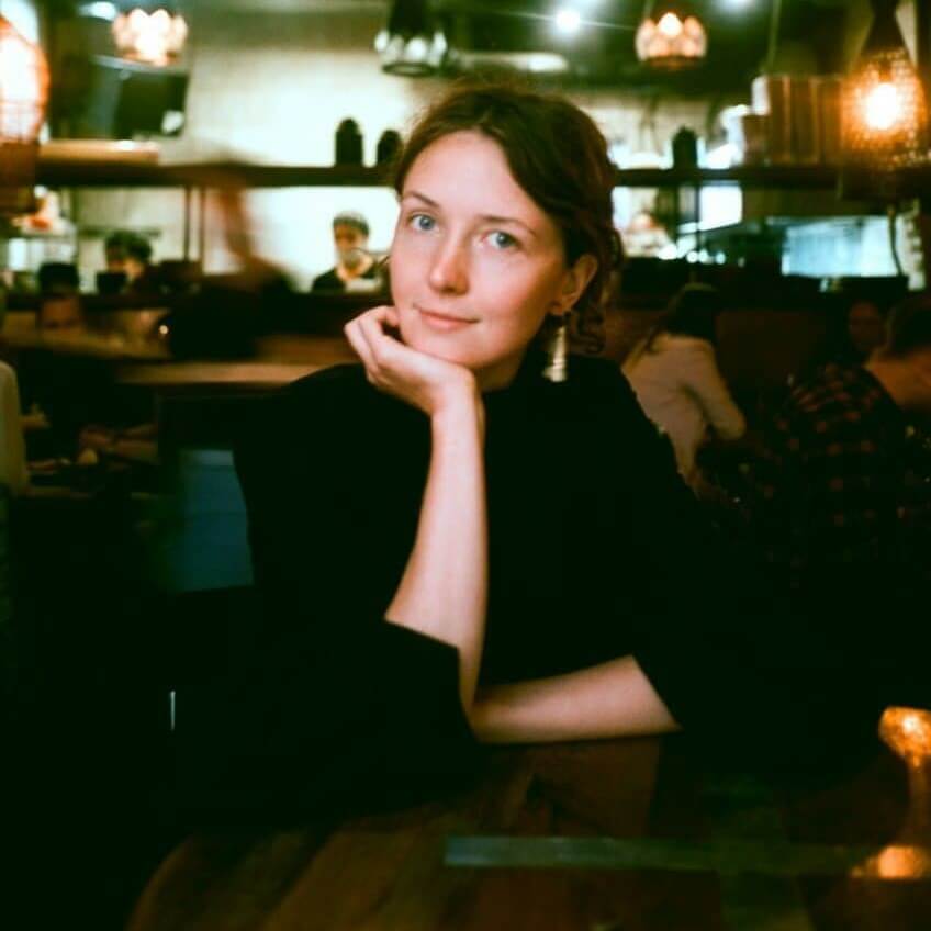 Портретная фотография женщины с каштановыми вьющимися волосами, с легким макияжем и серьгами, в черной кофте с длинными рукавами, сидящей за деревянным столом в помещении с приглушенным светом, подпирающей подбородок рукой. На заднем плане видны размытые фигуры людей.