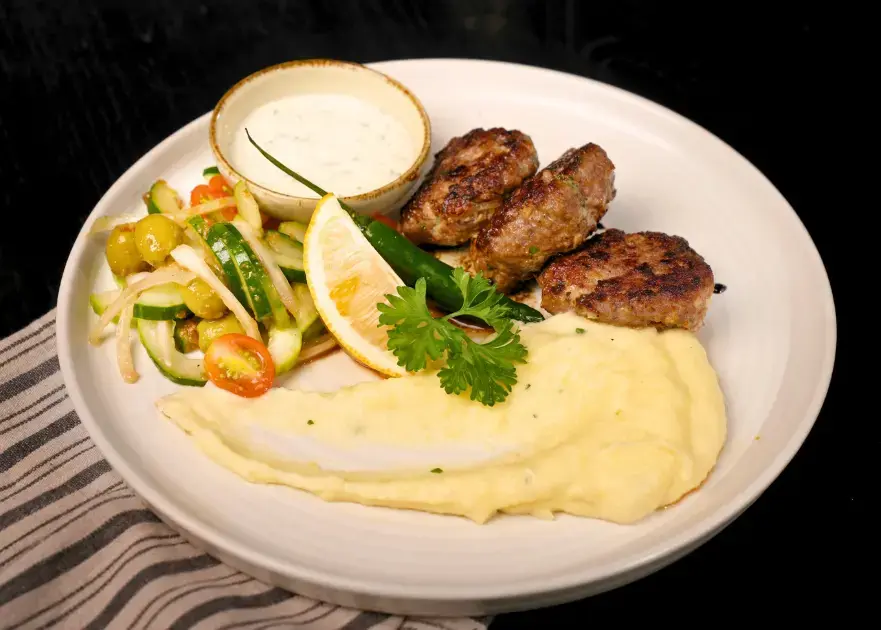 A beautifully plated dish of Mediterranean Bifteki featuring three grilled stuffed beef medallions, creamy mashed potatoes, a side salad, and tzatziki sauce—a hearty option for your Iftar in Ubud meal.