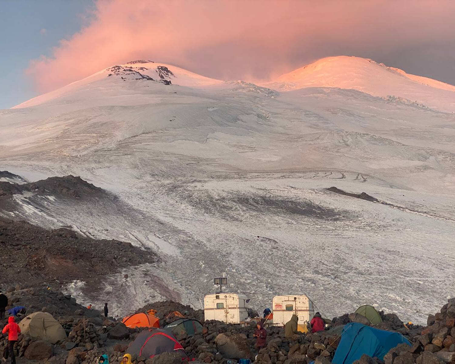 Elbrus summits glowing pink at sunrise above a camp of small cabins and tents