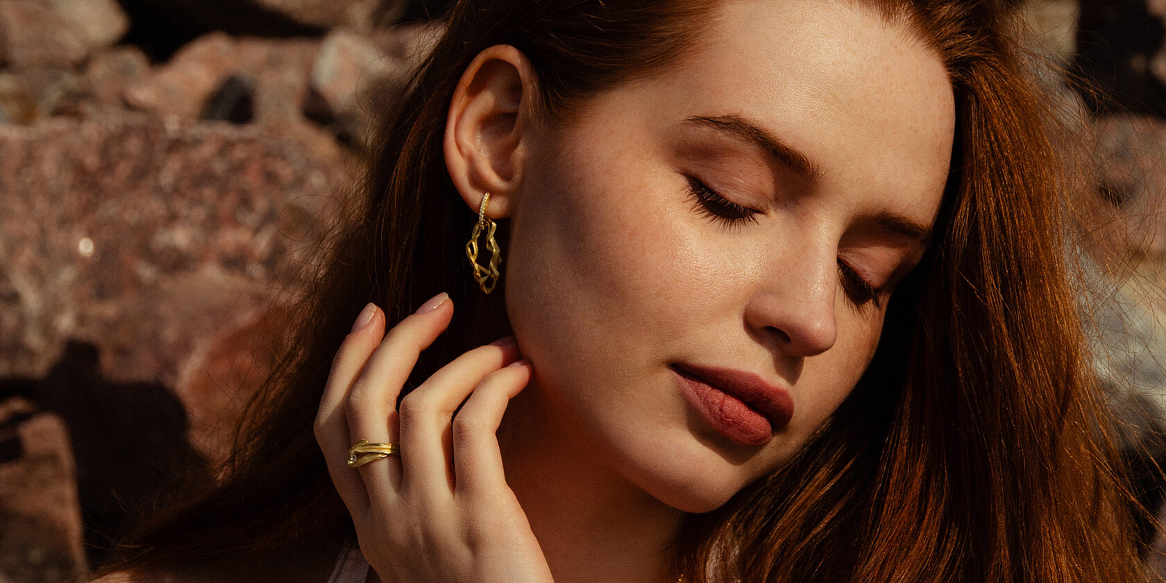 Девушка с рыжими волосами с золотыми украшениями сережками с переплетением деталей и бриллиантами в ушах на заливе на море в теплых тонах