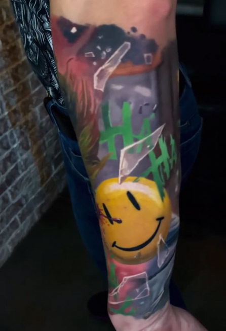 A colorful forearm tattoo in neo-traditional and realistic style, combining elements of DC Comics. The design includes the classic yellow smiley face with a blood trail (Watchmen), green 