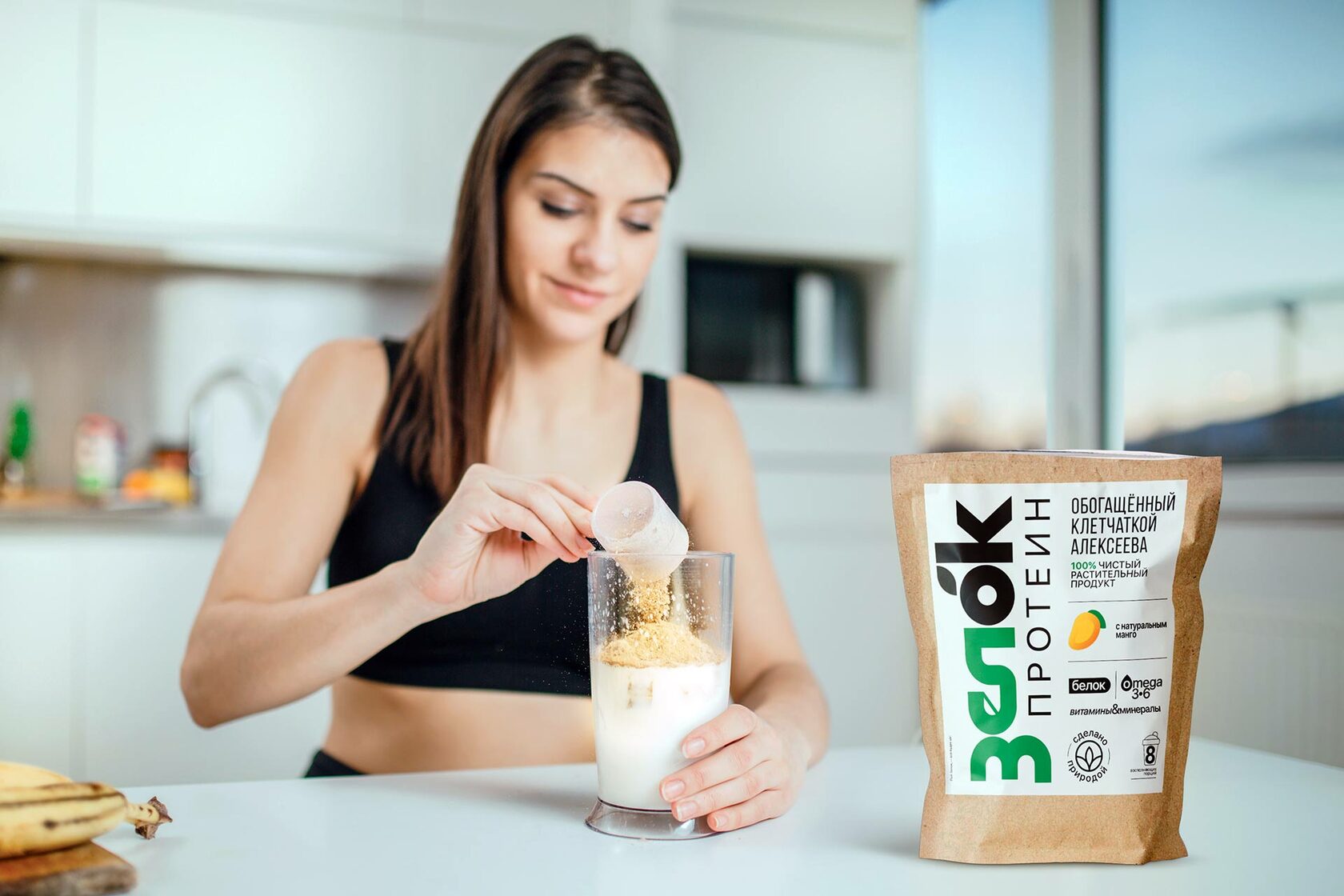 Красивая девушка стоит за кухонным столом, и готовит коктейль, насыпая растительный протеин в прозрачный шейкер. 