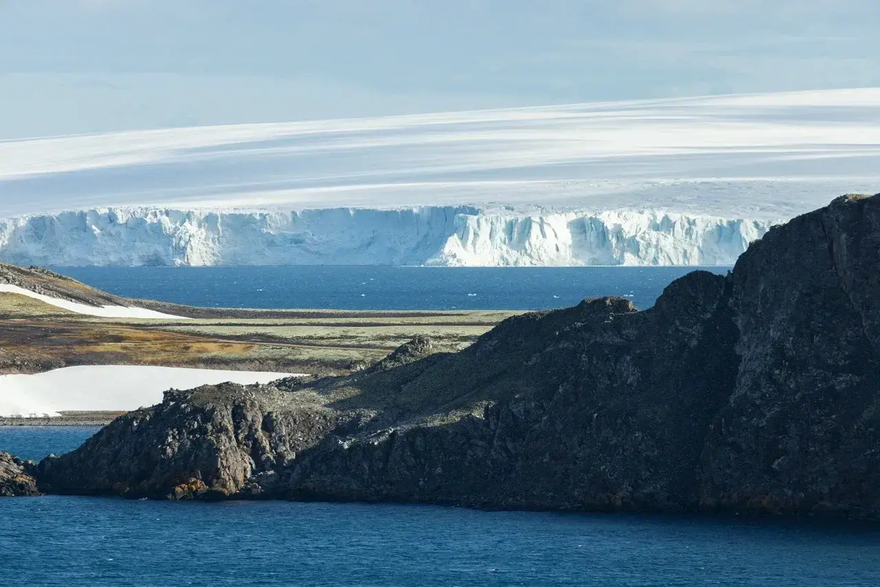 Dramatic Arctic landscape featuring dark rocky cliffs and a deep-blue bay in the foreground with a towering glacier wall and a vast ice sheet stretching across the horizon under soft polar light.