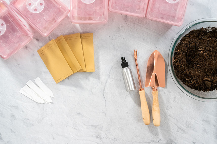 Gardening tools and supplies including seed packets, pink seed trays, a spray bottle, a copper trowel, and a bowl of soil, arranged on a light surface, illustrating essential items for seed starting and gardening practices.