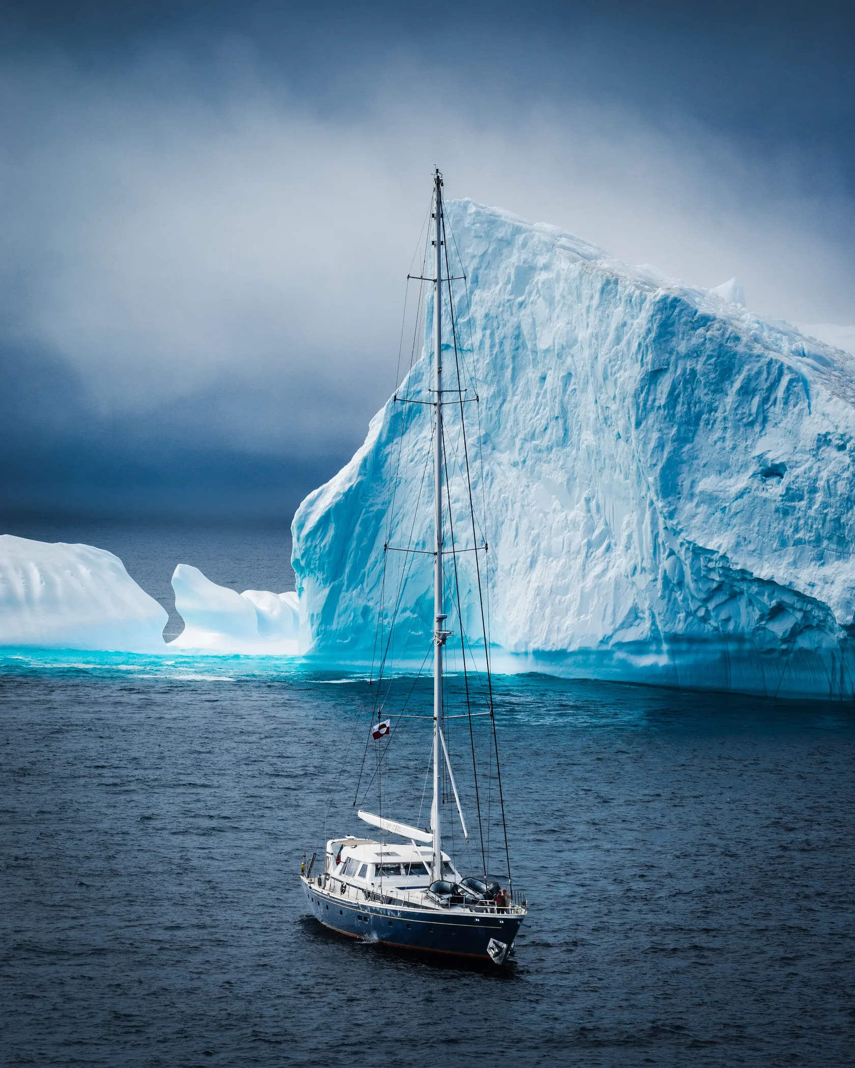 A sailing yacht navigating beside a towering blue iceberg in East Greenland. Massive Arctic icebergs drift through remote fjords, creating unforgettable landscapes for small-group sailing expeditions. Exploring Greenland by yacht offers close encounters with glaciers, pristine wilderness and some of the largest ice formations in the Northern Hemisphere