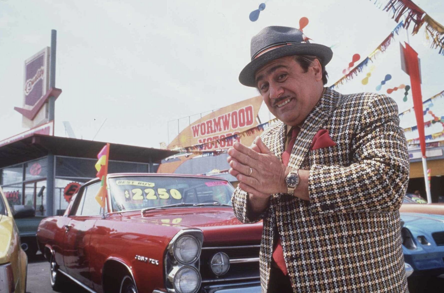 A dodgy-looking car salesman rubbing his hands together on the car lot