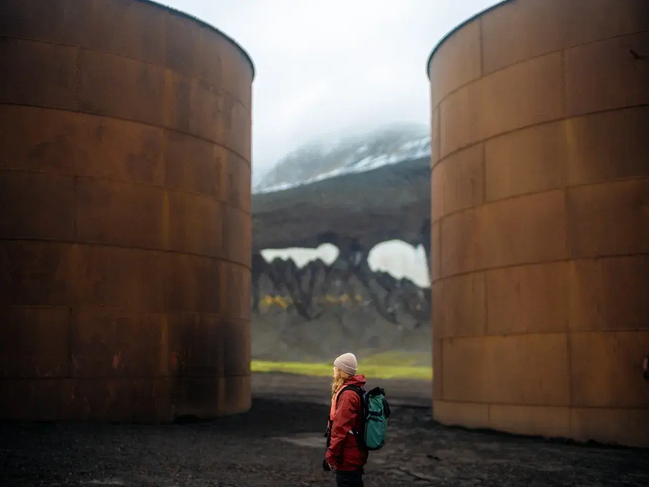 A traveler stands between two massive rusted fuel storage tanks on Deception Island, Antarctica, one of the continent's most unique volcanic landscapes. These abandoned tanks are remnants of the island's former whaling station, while volcanic mountains and black volcanic terrain create a dramatic backdrop, reflecting Deception Island's fascinating history and geothermal origins.