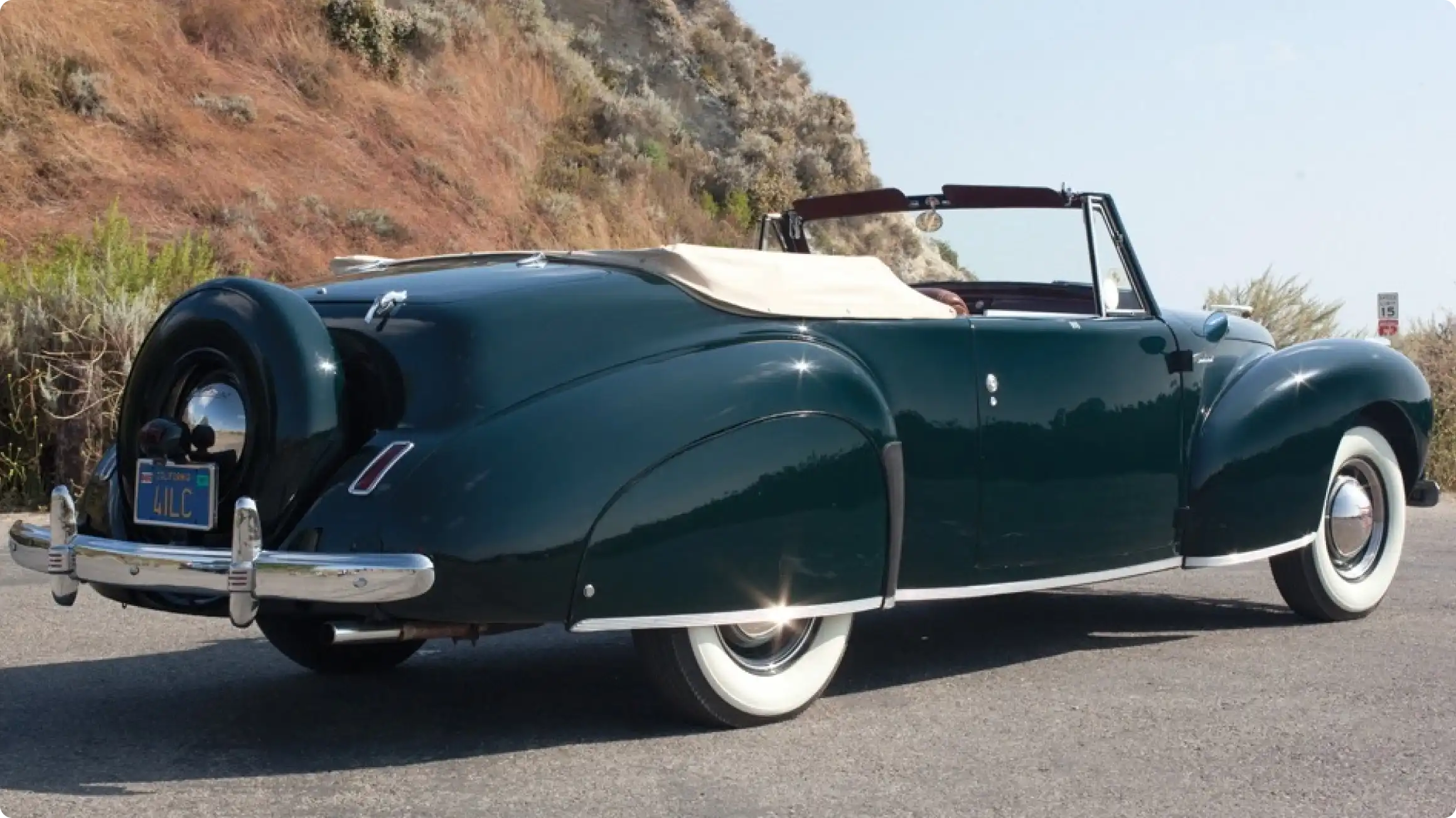 Классический кабриолет Lincoln Continental 1940-х годов в темно-зеленом цвете с белым верхом, припаркованный на шоссе на фоне холмов.