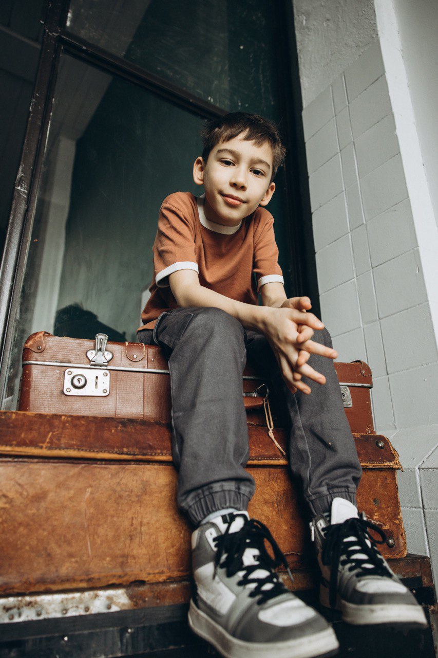 мальчик подросток сидит на чемоданах в коричневой футболке и кедах