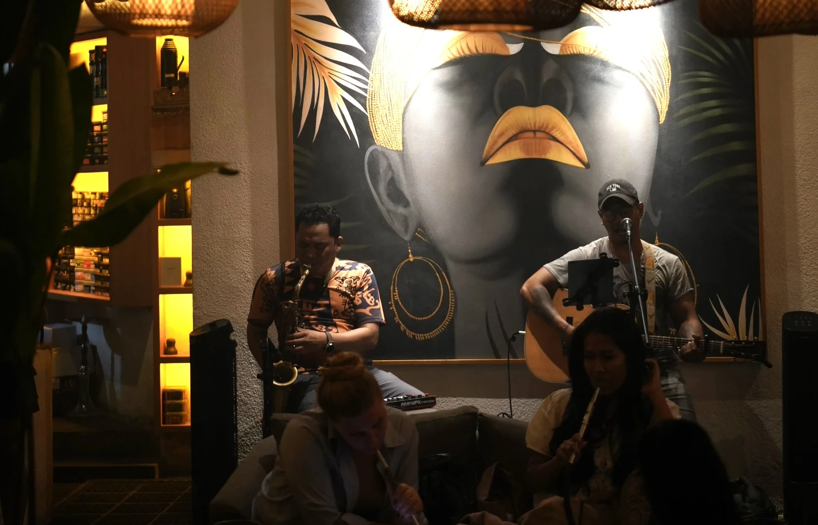 A live saxophone player and guitarist perform on a cozy stage in front of a distinctive face mural at Eden Hookah Club in Canggu, illustrating where you go on Wednesday in Bali for music.