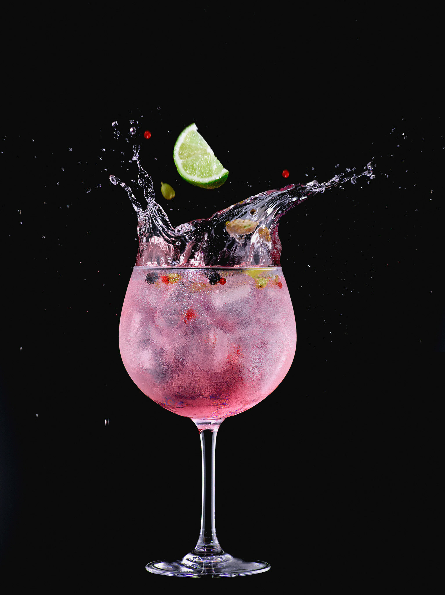 Изображение на чёрном фоне показывает винный стакан, в котором приготовлен коктейль &quot;джин-тоник малина&quot;. В стакане видны кубики льда ярко-розового цвета, а также долька лимона, которая лежит на краю стакана и выполняет функцию украшения. Коктейль заполнен ярко-красной жидкостью, которая поднимается к верху стакана и разбрызгивается, создавая впечатление движения. Стакан запотевший, что добавляет атмосферности и ощущения прохлады напитка.
