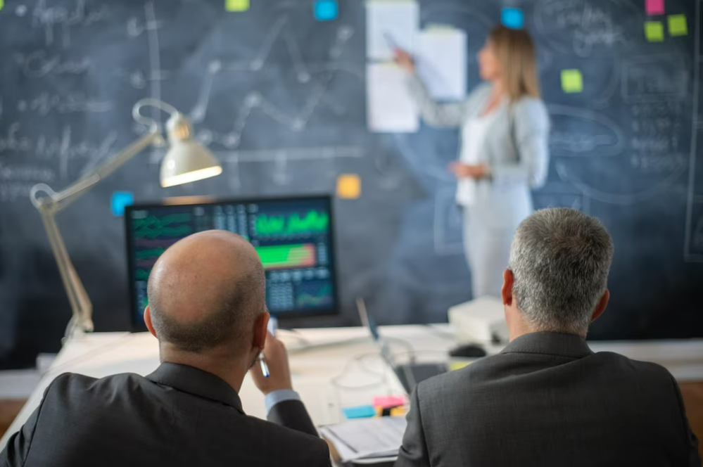 Dos hombres de espaldas tomando una clase para aprender trading. Observan a una mujer que señala gráficos en una pizarra