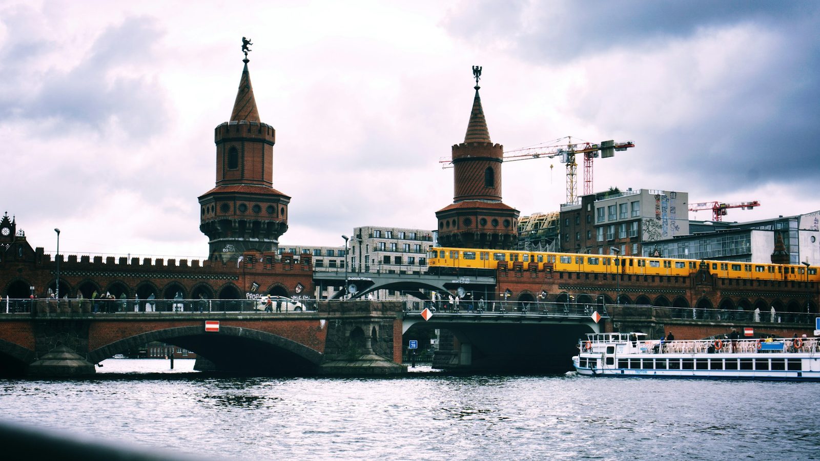 The red-brick Oberbaum Bridge over the River Spree in Berlin, with a yellow U-Bahn train crossing the upper deck and a tour boat below