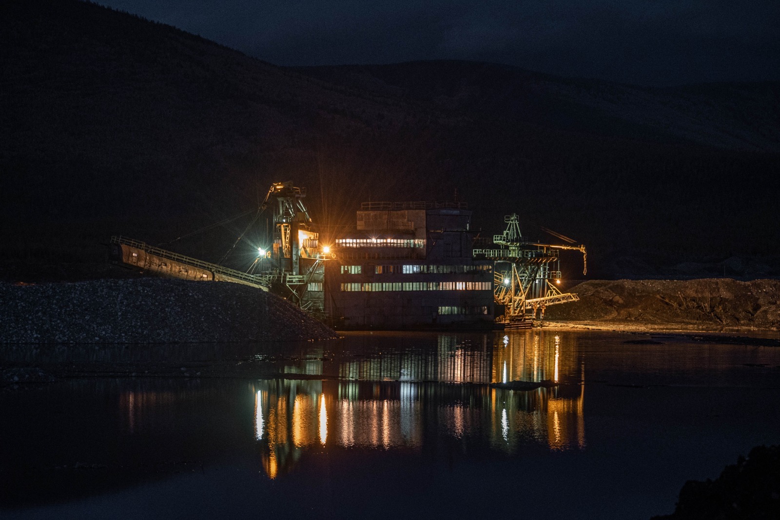 Дражный комплекс на участке золотодобычи ночью: работающая драга с подсветкой и отражением в воде, северный промышленный пейзаж