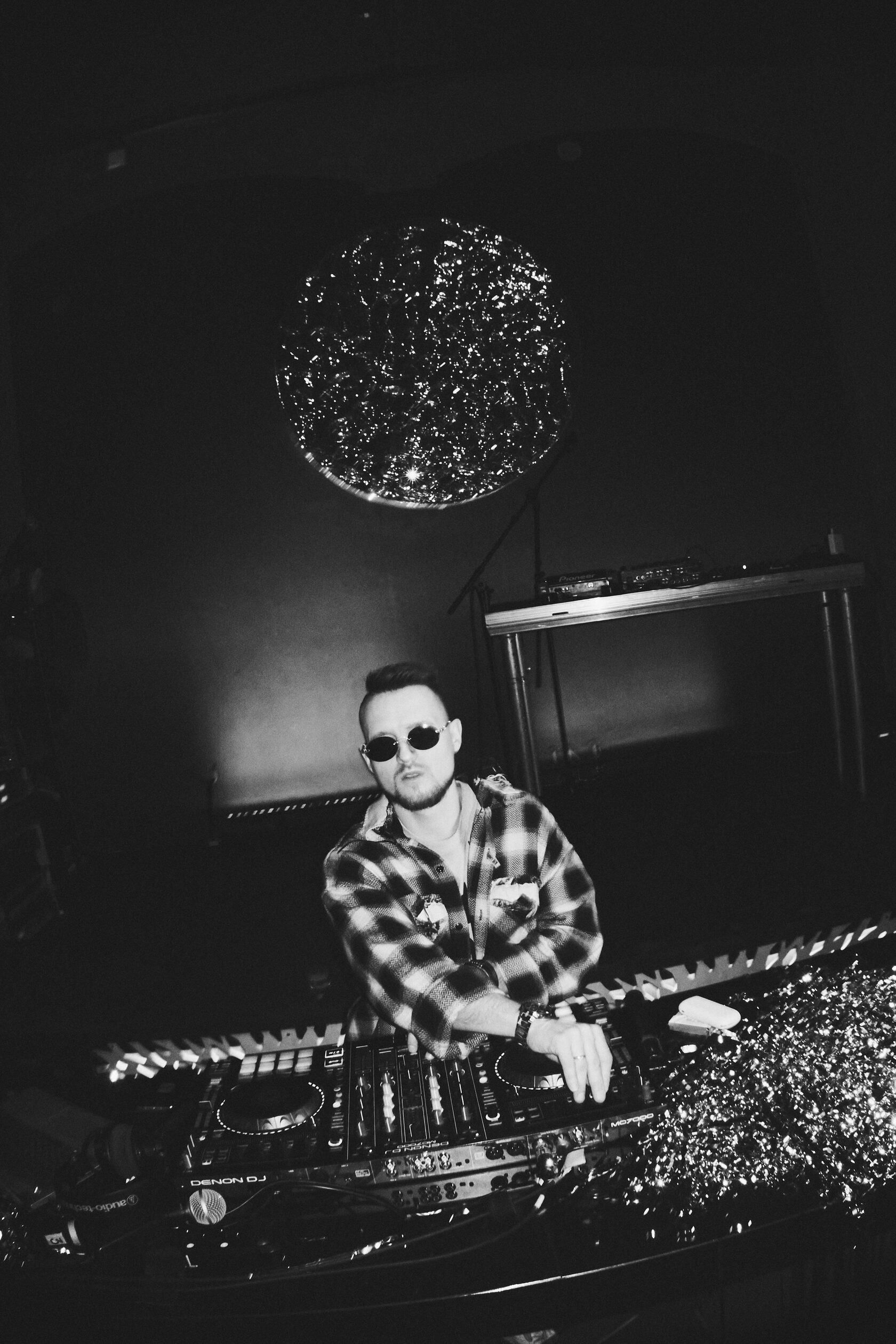 Чёрно-белый кадр DJ ROZHKOV в клетчатой рубашке за пультом под зеркальным шаром.
