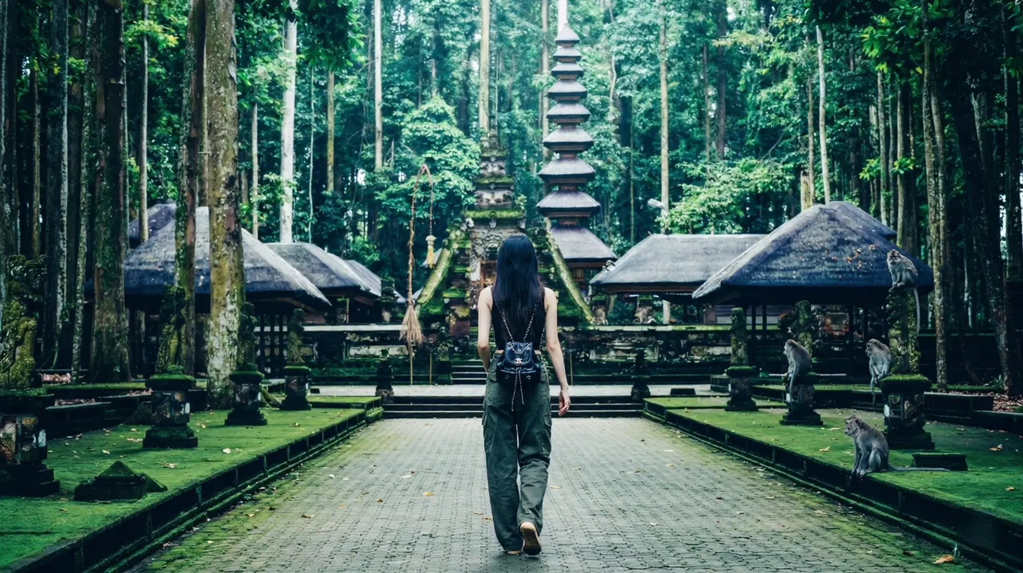A woman with a backpack walking along a scenic, moss-lined stone path toward a historic Balinese temple inside Monkey Forest Ubud.