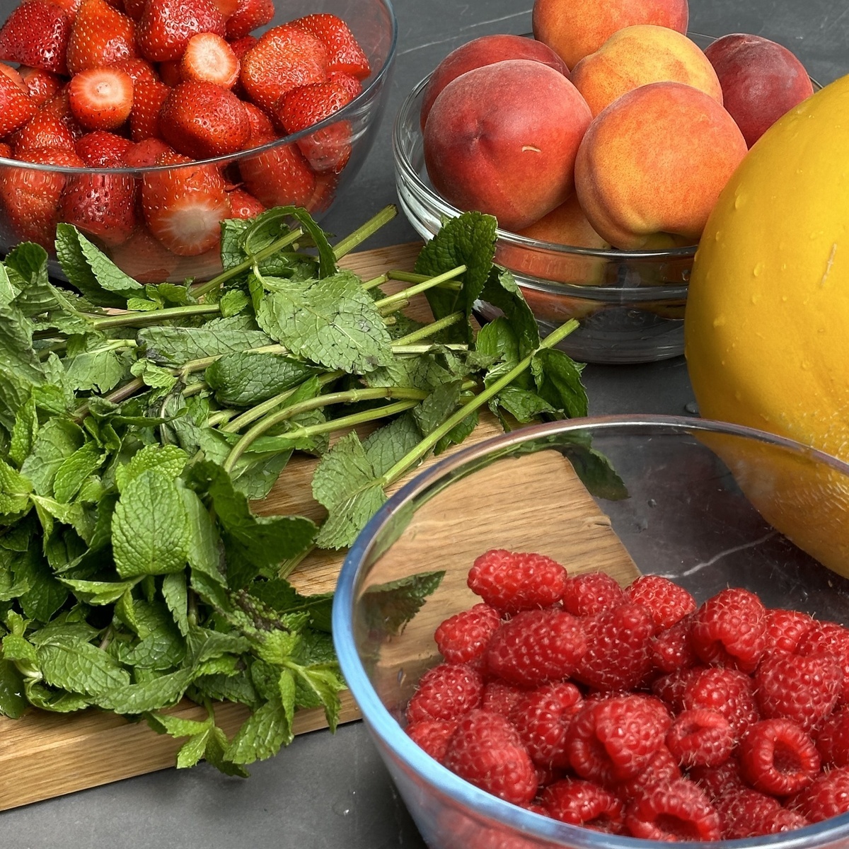 Изображение свежих ингредиентов для приготовления зимнего чая. На фото: клубника, персики, мята и малина, все красиво расположено в мисках и на деревянной доске, готовое к использованию в рецептах для ароматных напитков.