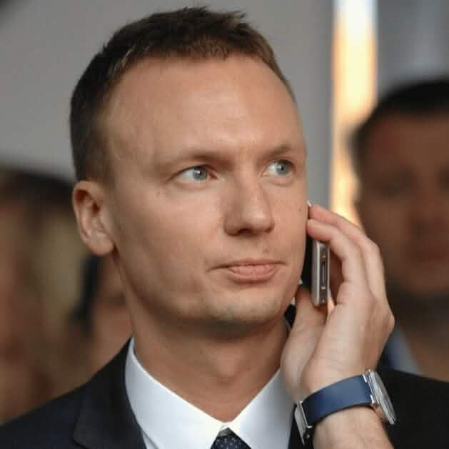 Портретная фотография мужчины с короткими светлыми волосами, в темном деловом костюме, белой рубашке и синем галстуке с белой полосой, держащего в руке мобильный телефон и смотрящего влево с невозмутимым выражением лица. На заднем плане видны размытые силуэты других людей.