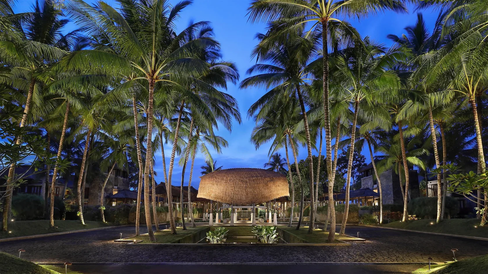 Beautifully illuminated palm tree-lined entrance and striking bamboo architecture at Maya Ubud Resort & Spa at twilight.