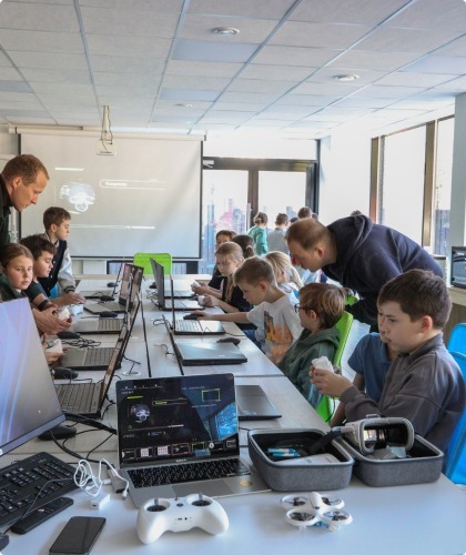 Дети на занятии по робототехнике и программированию сидят за столами с ноутбуками, педагог помогает им работать с оборудованием и контроллерами. На переднем плане — ноутбук, геймпад и кейс с электроникой.
