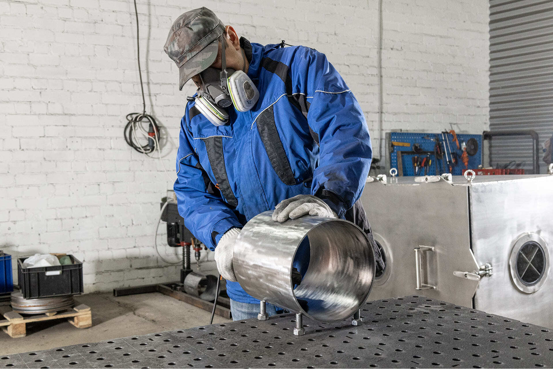 Сотрудник в защитной маске и перчатках обрабатывает цилиндрический элемент вакуумной камеры из нержавеющей стали в производственном цехе.