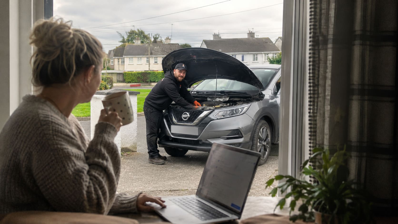 Woman working at home with a coffee while her car is being serviced outside by a mobile mechanic