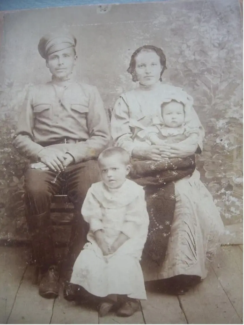 Архивная чёрно-белая фотография начала XX века: молодой мужчина в военной форме сидит рядом с женщиной, держащей на руках младенца; перед ними стоит маленькая девочка в светлом платье. Семья запечатлена в фотостудии на фоне декоративной ширмы.