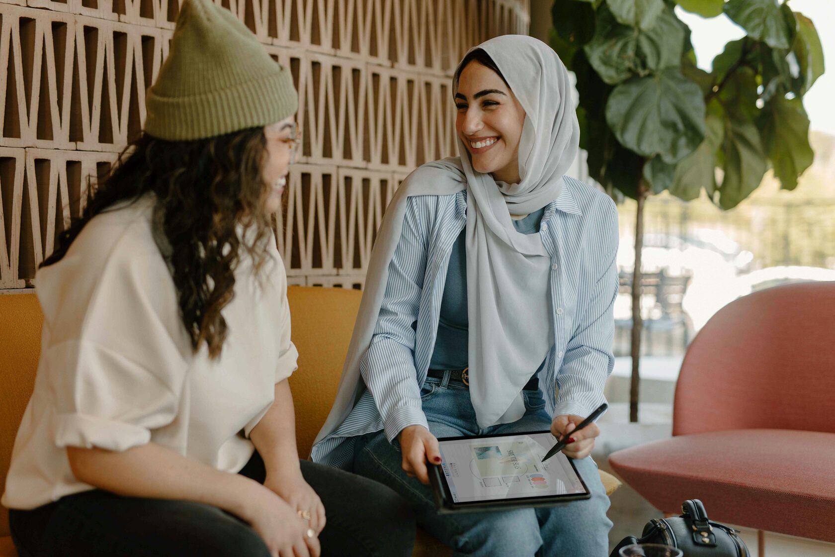 Two women wearing hijab sitting on a couch, using a tablet.