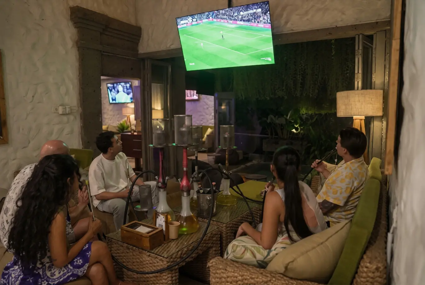 A high-angle view of a group of friends relaxing comfortably on plush sofas, sharing several multi-hose hookahs, and watching a live football match together on a large wall-mounted screen. Combining premium shisha with big sports games is the best event i