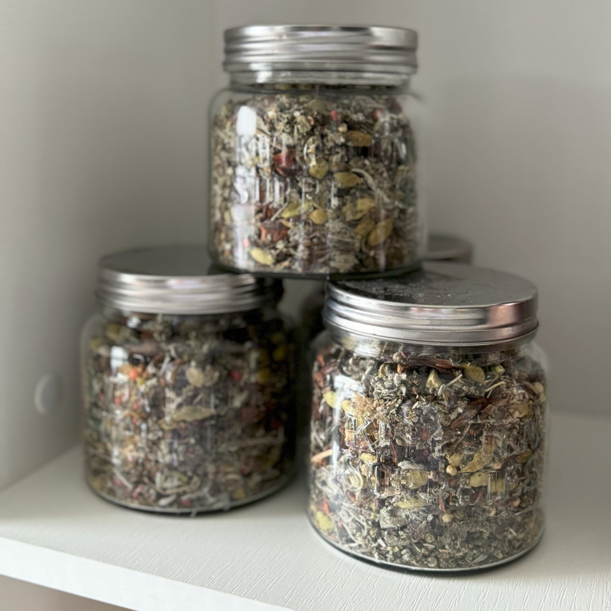 Изображение трех стеклянных банок с травяными заготовками для чая. Банки содержат смесь сушеных трав и специй, таких как мята, чабрец и другие растения, используемые для приготовления ароматного чая.