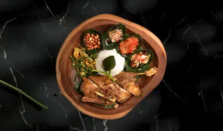 A top-down view of a rustic wooden plate serving crispy fried duck alongside white rice, Balinese green bean salad, roasted peanuts, and three sambals. A comforting local favorite to break the fast.