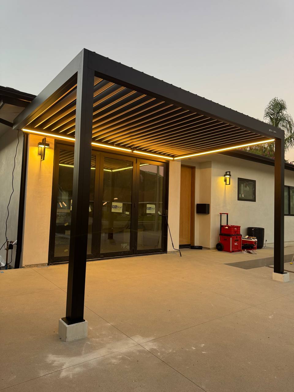 Warm LED strips illuminate the louvered roof and concrete patio beside the house.