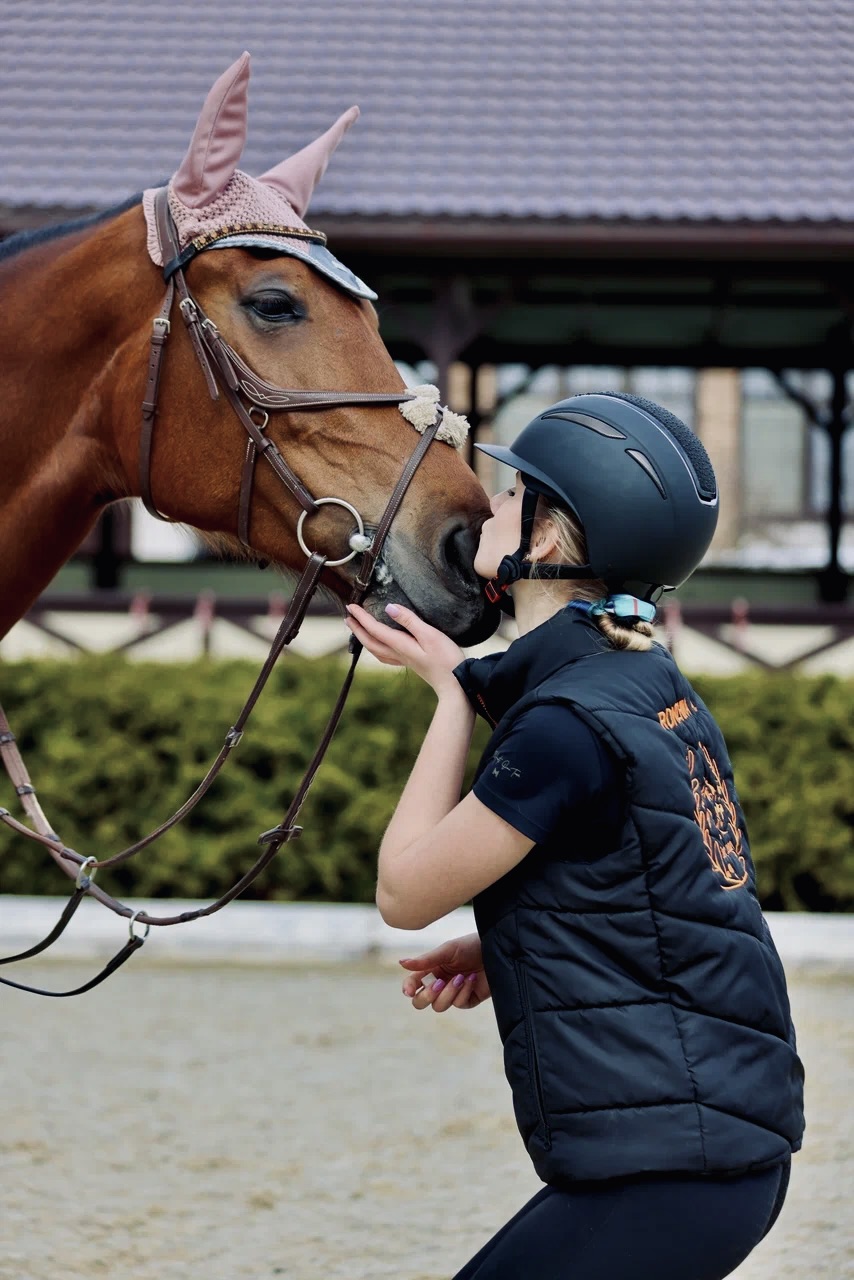 Девушка в шлеме нежно целует морду лошади, стоя рядом с ней на тренировочной площадке
