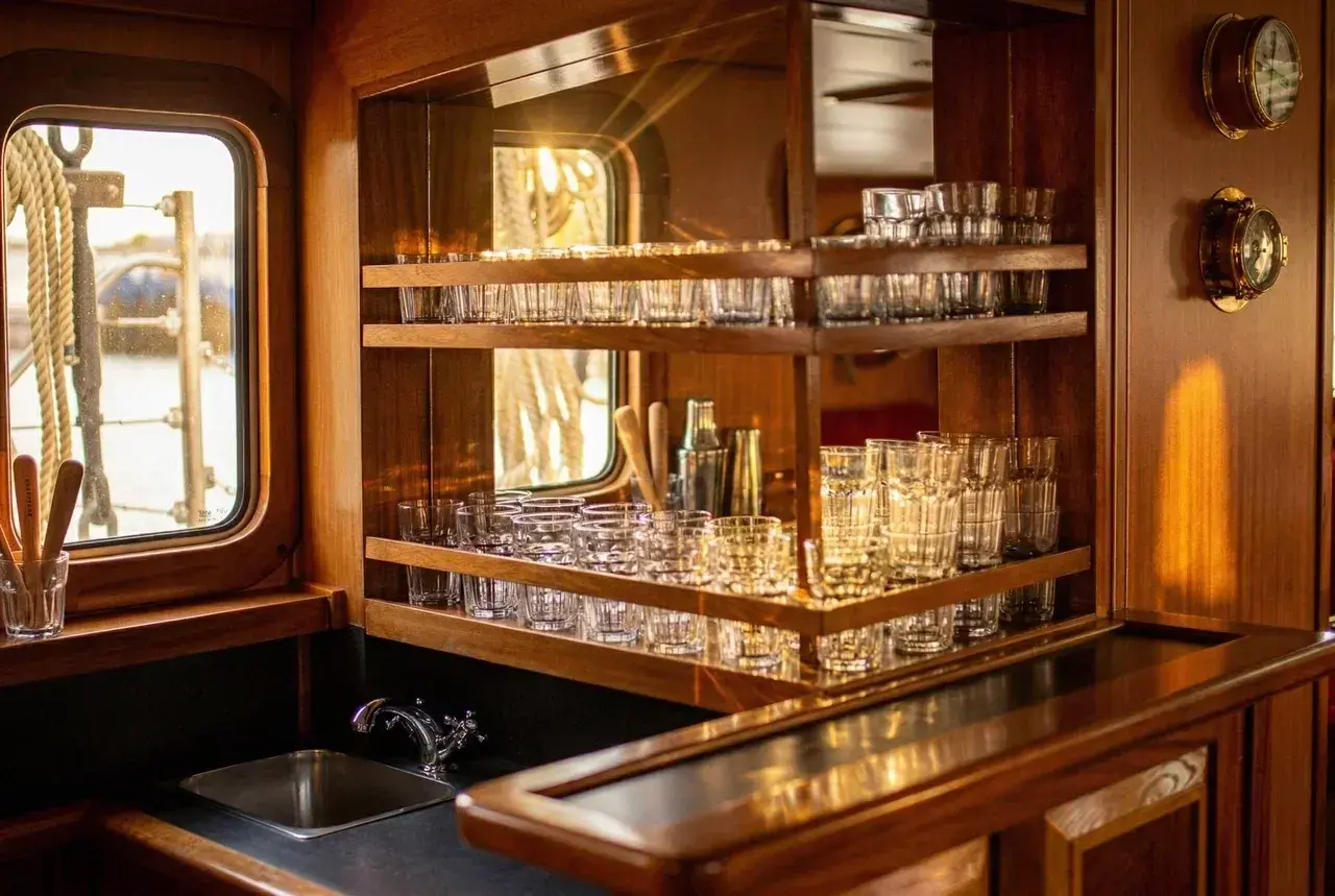 Warm wooden interior of the schooner Eldorado, featuring a beautifully crafted onboard bar with shelves of glassware illuminated by golden sunlight streaming through porthole windows. Nautical instruments and polished wood details create a cozy and elegant atmosphere, highlighting the comfort of life aboard the expedition vessel.