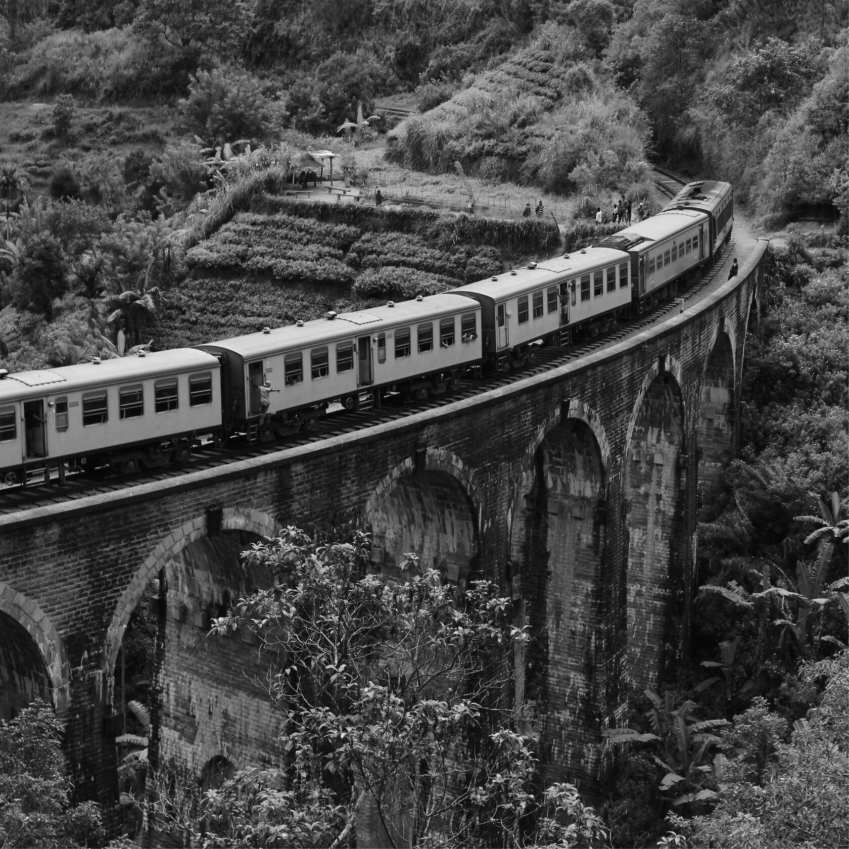Чёрно-белая фотография гигантской статуи бога Муругана у входа в пещеры Батур (Batu Caves) в Малайзии. На заднем плане видна лестница, ведущая к пещерам, окружённая густой растительностью и скальным массивом. Монументальная фигура статуи доминирует в композиции, подчёркивая религиозное и культурное значение этого места. 