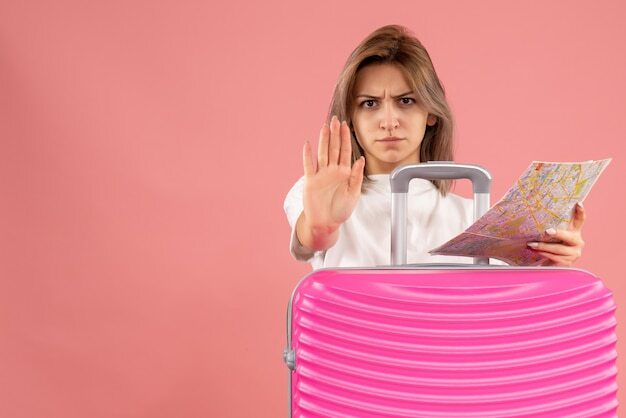 Front view of dissatisfied young girl with pink suitcase holding map making stop sign