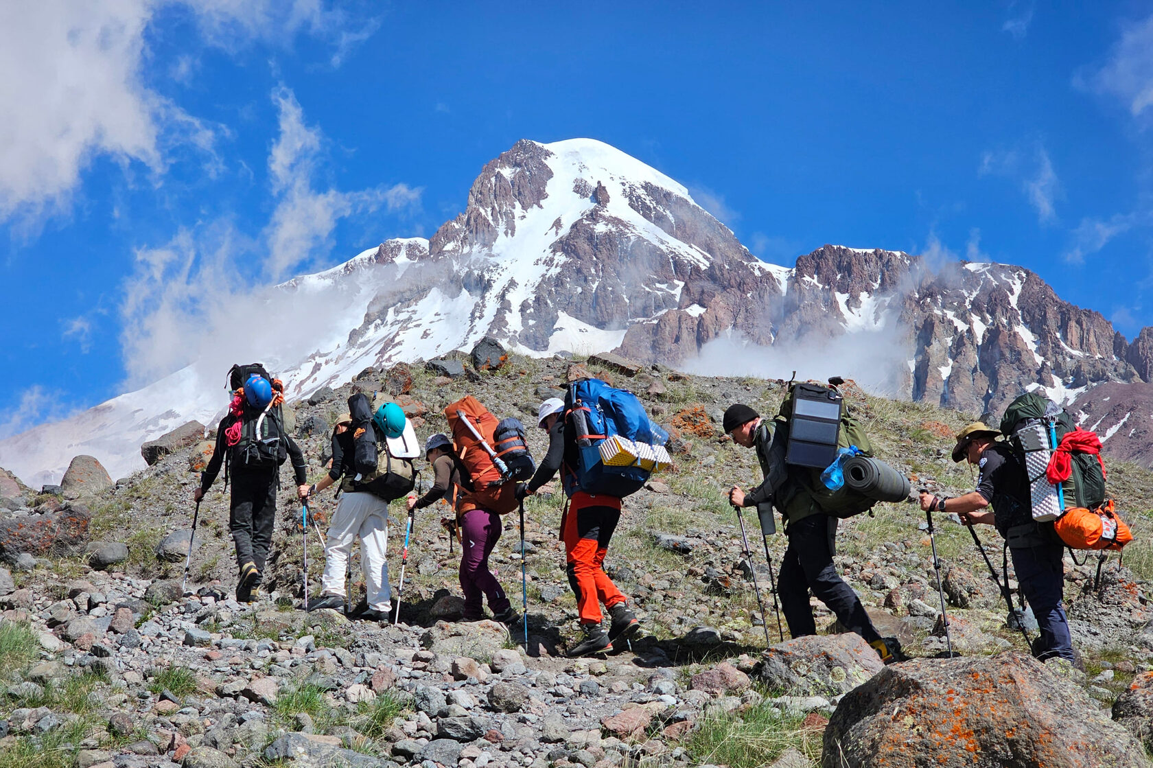 Trekkers with backpacks heading up a rocky slope towards snow-capped Kazbek