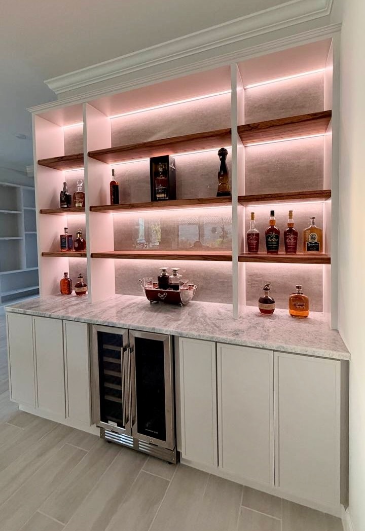 Custom built-in bar cabinetry with warm LED shelf lighting, natural wood shelves, dual beverage refrigerators, and decorative crown molding